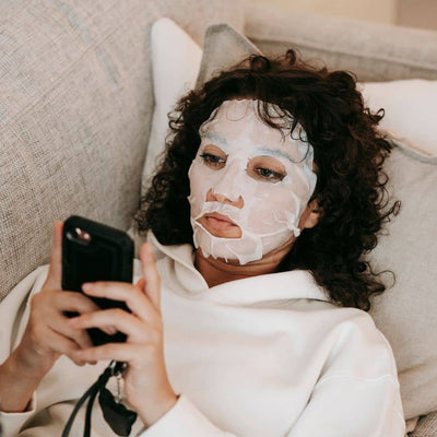 woman laying on sofa, using cellphone while wearing a face mask.
